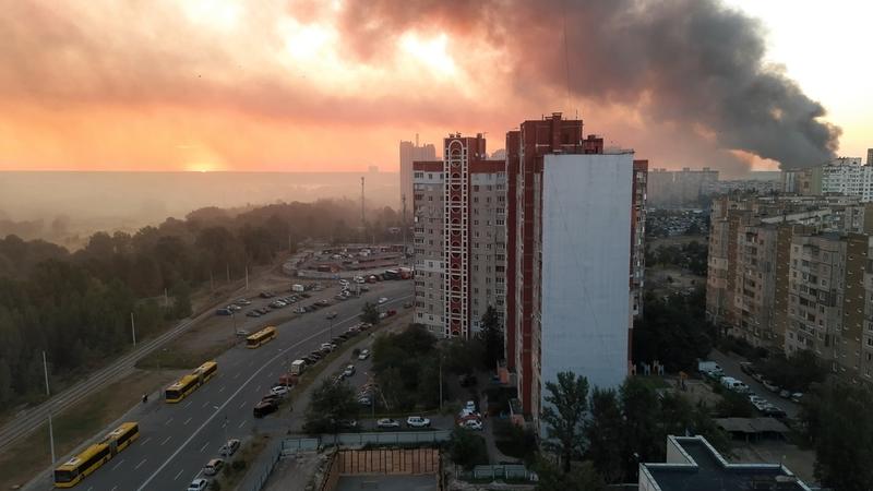 Smoke rises from buildings in a cityscape at sunset, with orange skies and a road lined with yellow buses.