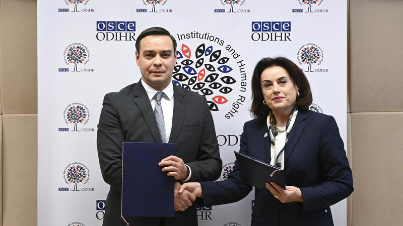 Two people in formal attire shake hands while holding folders, standing in front of a branded backdrop.
